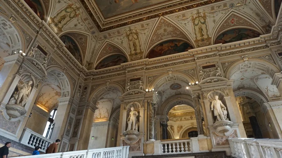 Ornate grand staircase with marble statues, arches, and intricate ceiling frescoes in a historic building.