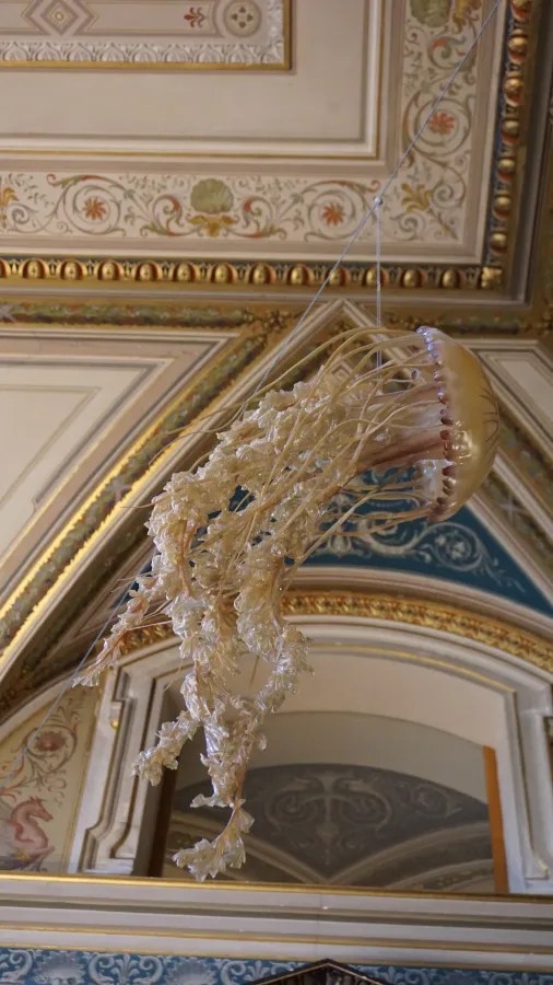 A realistic jellyfish sculpture suspended from a grand, ornately decorated and gilded ceiling.
