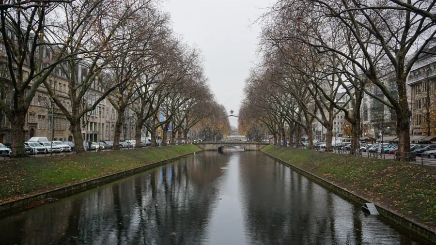 Tree-lined canal reflecting leafless trees and a bridge under an overcast city sky.