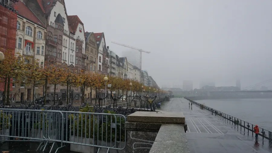 Historic buildings along a foggy riverfront promenade with autumn trees on a gray day