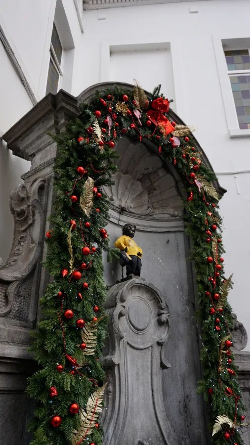 Manneken Pis statue dressed in yellow, surrounded by a festive evergreen garland with red baubles.