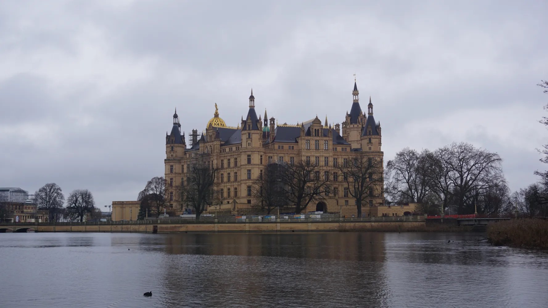 Grand multi-towered palace on a lakeside under grey clouds with reflections in the water.
