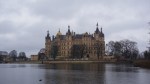 Grand multi-towered palace on a lakeside under grey clouds with reflections in the water.