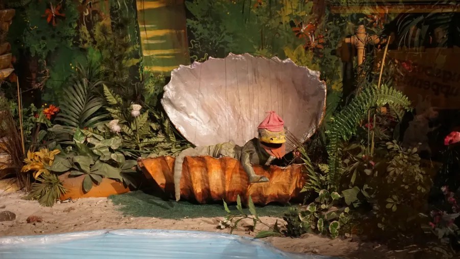 Puppet lizard wearing a pink hat inside a large clamshell in a jungle-themed display