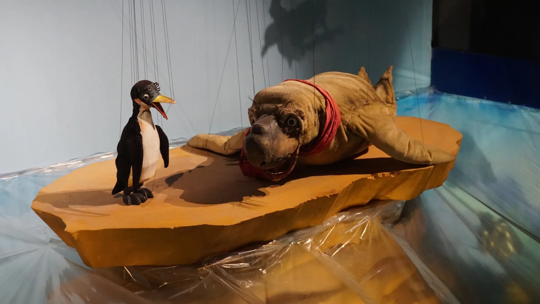 Penguin and walrus marionettes with a red scarf on a theatrical stage set.