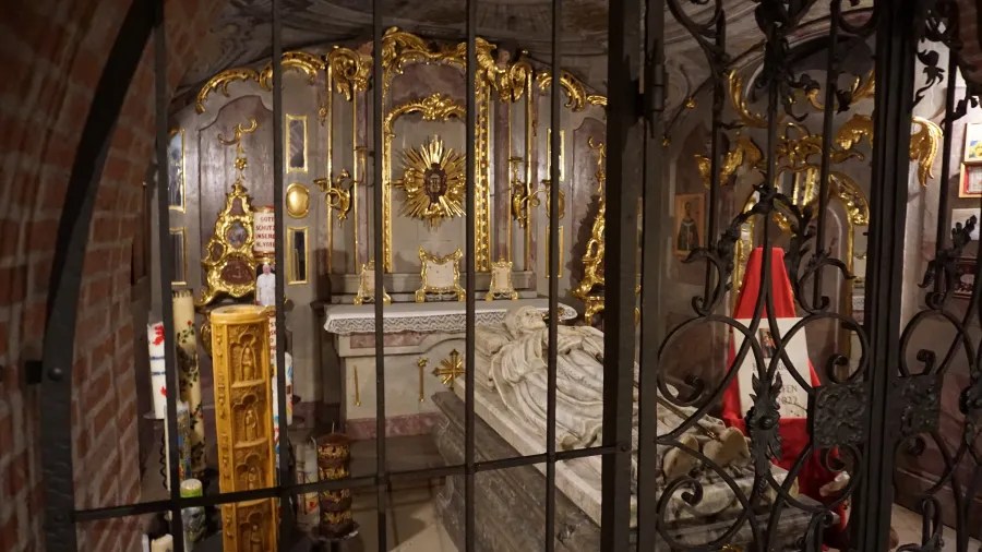 Ornate crypt featuring a stone sarcophagus and gold altar, viewed through a wrought-iron gate.