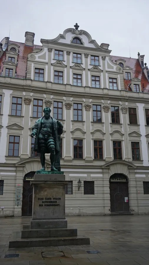 Statue of Hans Jakob Fugger before an ornate building. Text: HANS JAKOB FUGGER BEFÖRDERER DER WISSENSCHAFT, nine five.