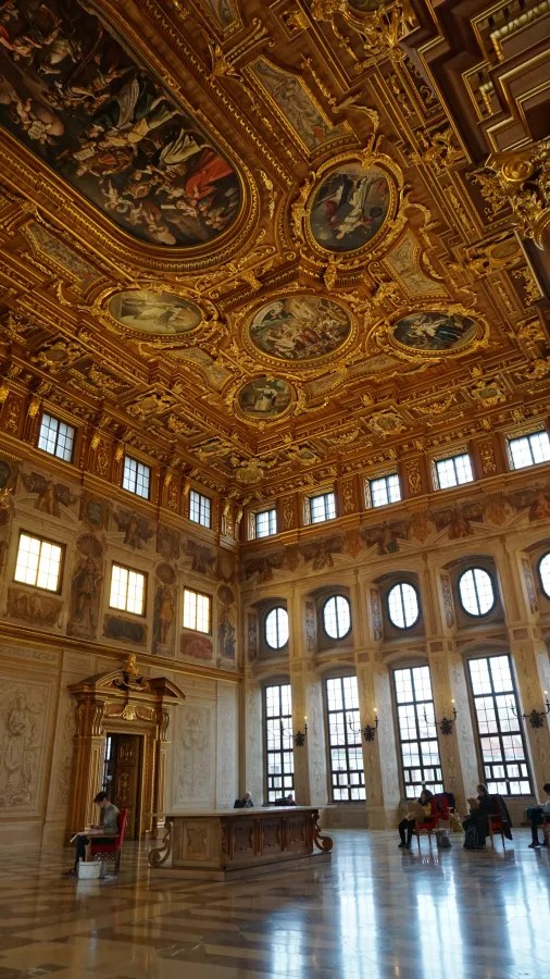 Grand hall featuring an intricate gold ceiling with paintings and tall, bright windows
