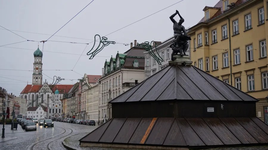 A rain-slicked city street featuring a covered fountain statue and a large domed church.