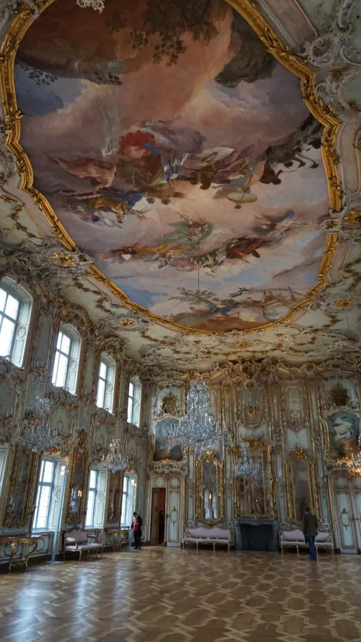 Grand hall featuring a large ceiling fresco, ornate gold-leaf decorations, and crystal chandeliers.
