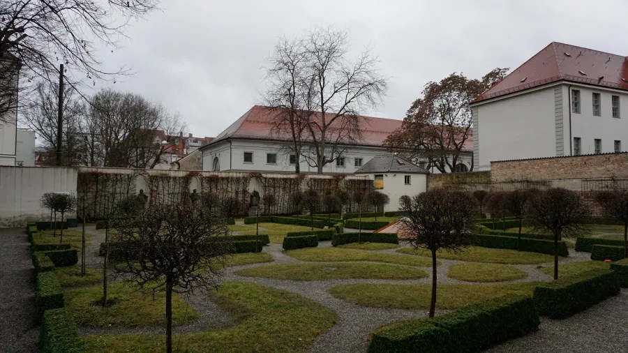 Formal garden with manicured hedges and small trees in front of buildings with red-tiled roofs.
