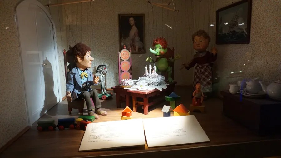 Marionette puppets in a detailed miniature room display with furniture, toys, and informational plaques.