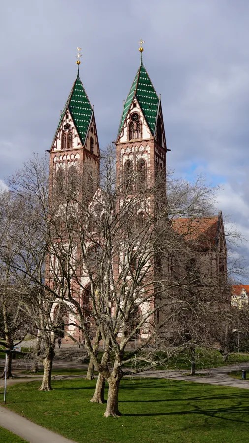 Reddish-brown stone church with twin green-tiled spires, partially obscured by bare trees in a park.