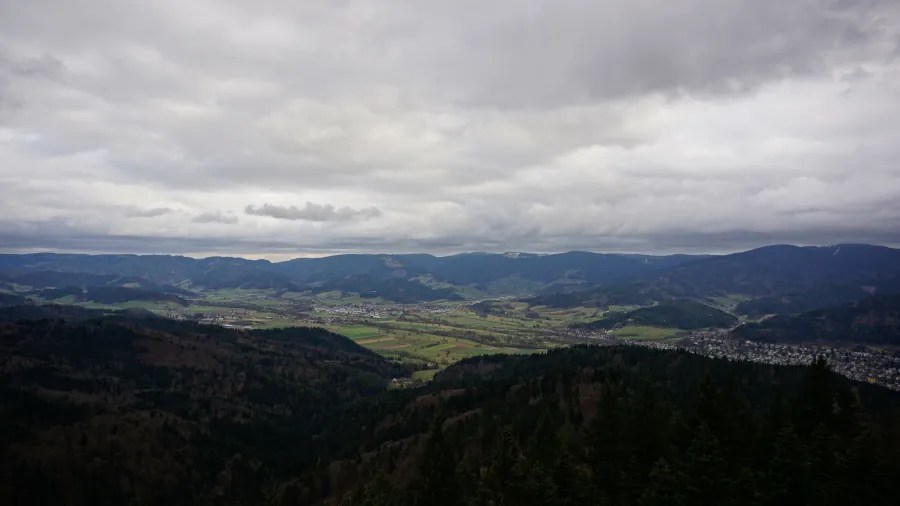Aerial view of a green valley with small towns surrounded by forested mountains under cloudy skies