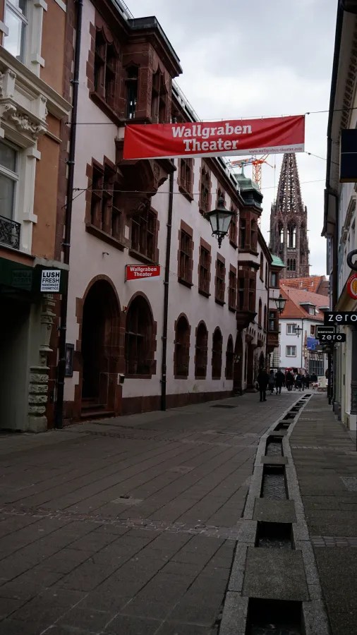 A narrow street with a Wallgraben Theater banner, a stone water channel, and a cathedral spire.