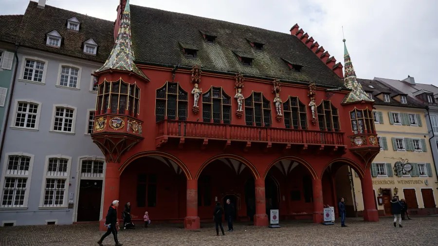Ornate red historic building with statues, turrets, and a balcony above an arched walkway.