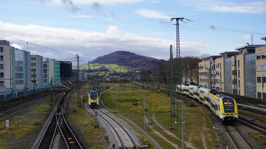 Yellow and white trains on railway tracks with buildings and a mountain in the background.
