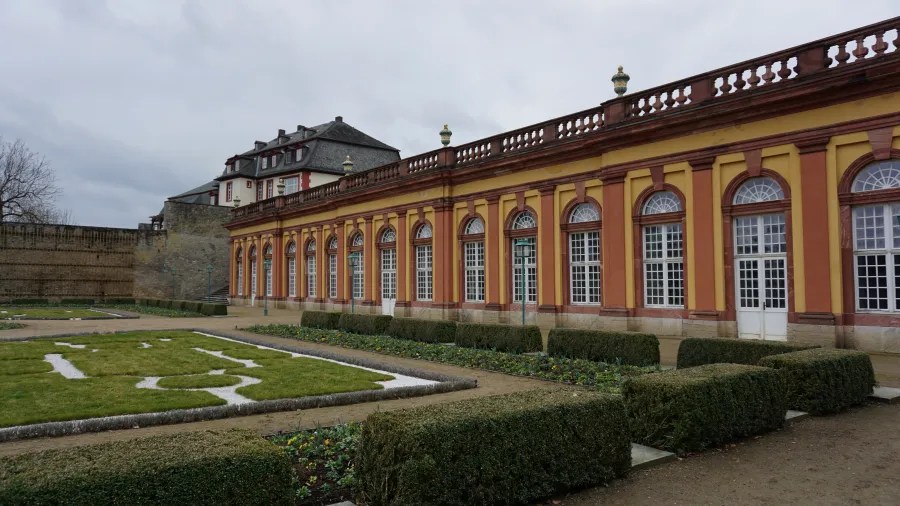 Long yellow building with arched windows and manicured formal gardens under a gray sky