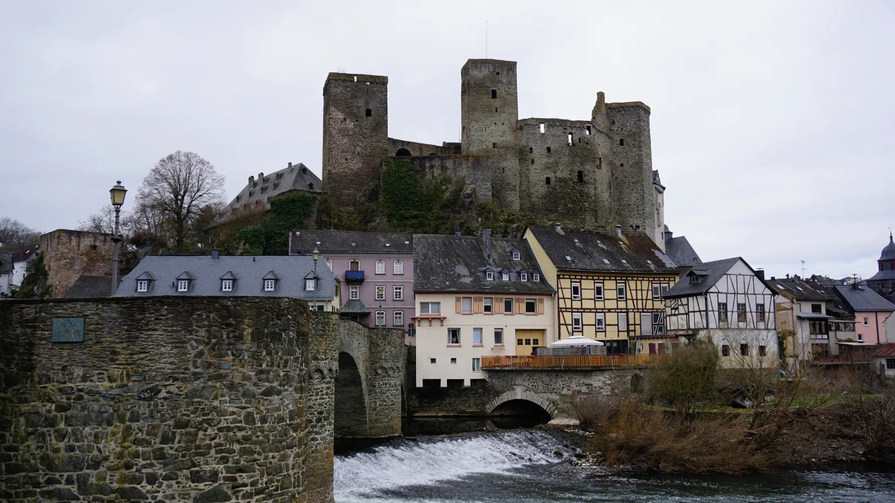 Medieval stone castle ruins overlooking colorful timber-framed houses and a river with a stone bridge.