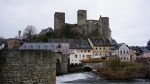 Medieval stone castle ruins overlooking colorful timber-framed houses and a river with a stone bridge.