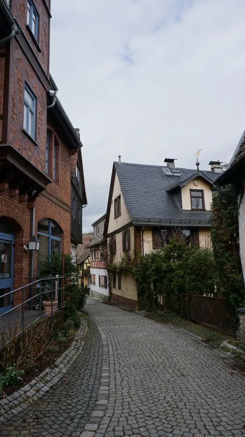20230319_Deutschland_Hessen_3-Burgen-Weg (12) A narrow cobblestone alleyway winding between traditional half-timbered and brick buildings.