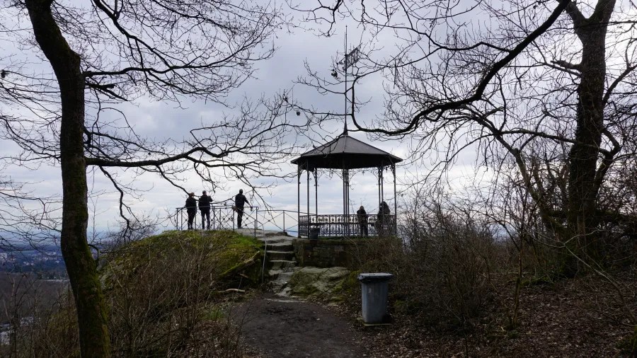 20230319_Deutschland_Hessen_3-Burgen-Weg (21) Silhouetted figures at a scenic hilltop lookout featuring a decorative metal gazebo and bare trees.