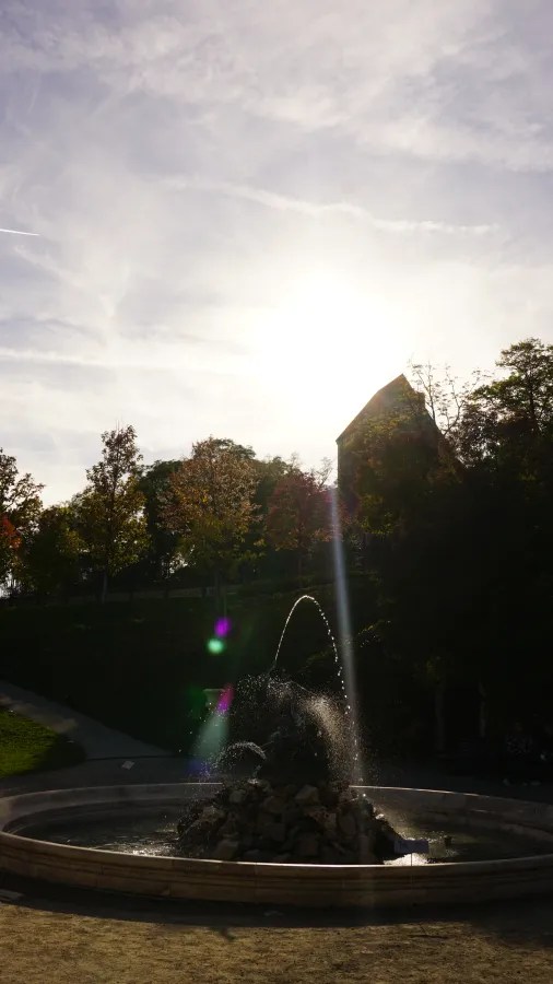 Stone fountain spraying water in a park with trees silhouetted against a bright sun.