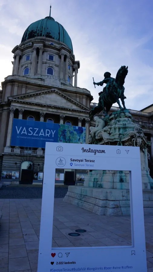 Buda Castle dome and Savoy monument behind an Instagram frame. Banner: VASZARY AZ ISMERETLEN ISMERŐS | REDISCOVERED. Frame: Instagram, Savoyai Terasz, 2.022 kedvelés, #SavoyaiTerasz #BudaiVár #longdrinks #beer #wine #coffees.