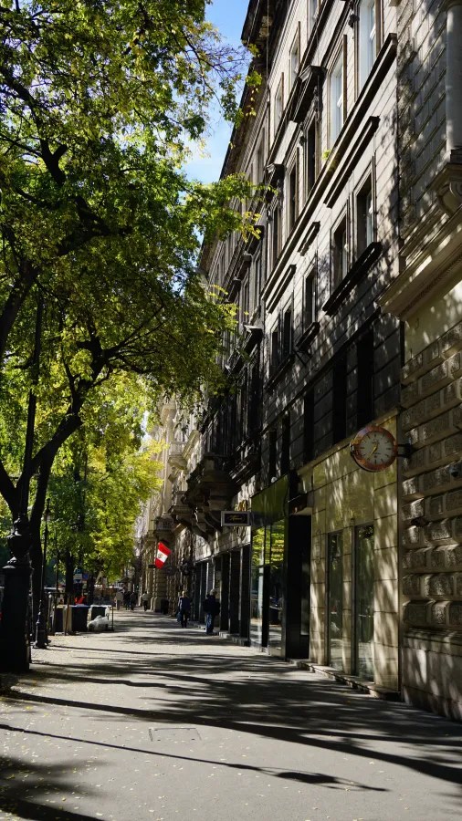 Tree-lined sidewalk beside historic European buildings with long shadows on a sunny day