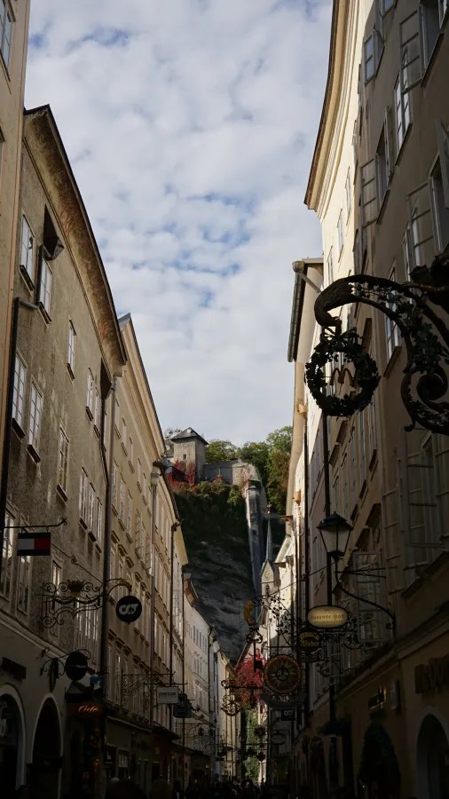 Narrow street with historic buildings and ornate signs leading to a fortress on a cliff