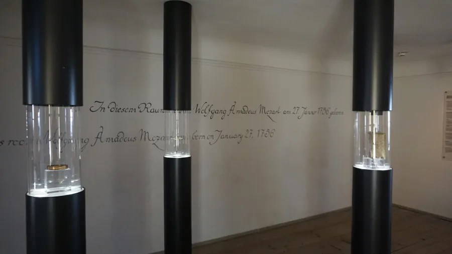 A museum room with black pillars containing glass display cases and wall text stating: In diesem Raum wurde Wolfgang Amadeus Mozart am 27. Jänner 1756 geboren. In this room Wolfgang Amadeus Mozart was born on January 27, 1756.