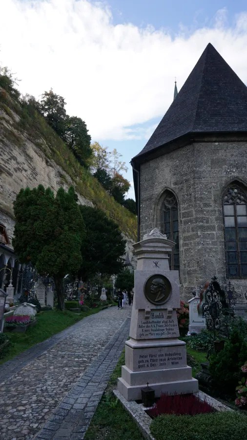 Cobblestone path through a historic cemetery with stone monuments, a chapel, and a steep rocky cliff.