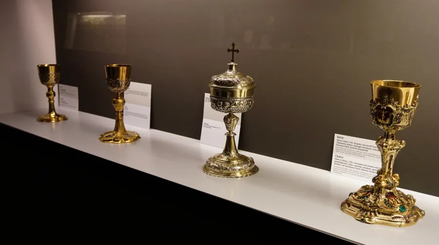 Four ornate gold religious chalices and a ciborium displayed on a museum shelf