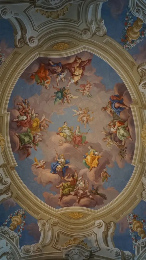 Baroque ceiling fresco depicting numerous figures and cherubs among clouds within an ornate oval frame.