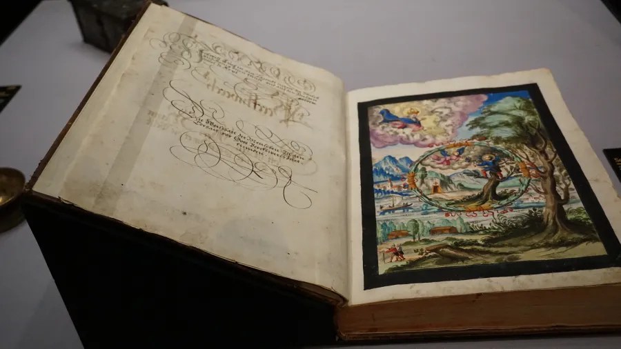 Open antique book with elaborate German calligraphy and a detailed landscape miniature illustration.