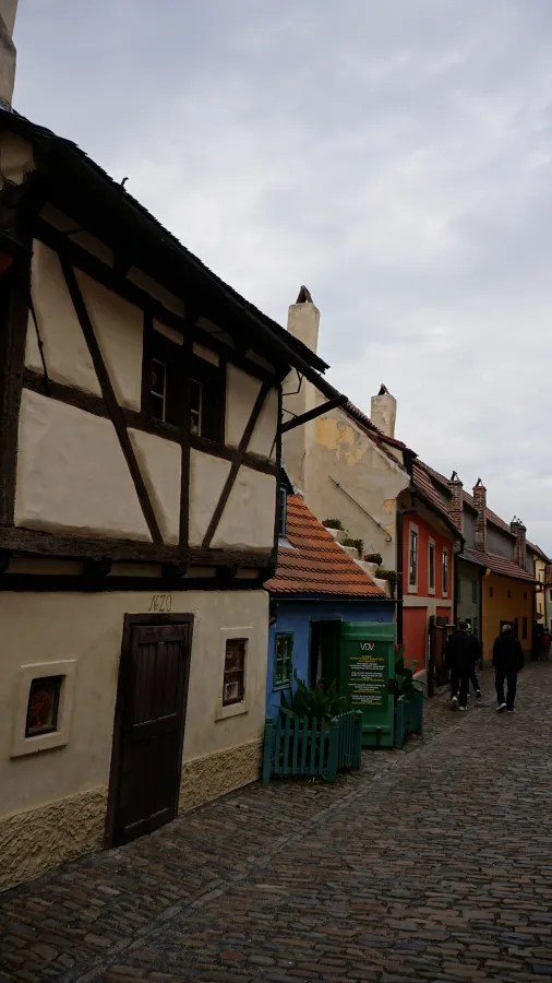 20221021_Tschechien_Prag (146) Small, colorful historic houses along a narrow cobblestone street under a cloudy sky.