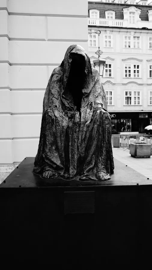 20221021_Tschechien_Prag (48) Bronze sculpture of a seated, cloaked figure with an empty face in a city square.