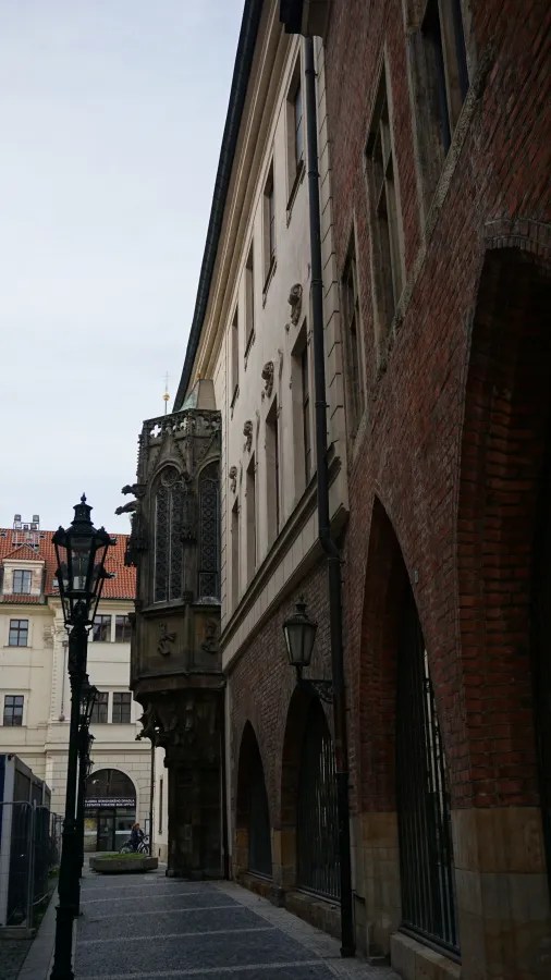 Narrow historic street featuring a Gothic stone oriel window and a red brick arched building.