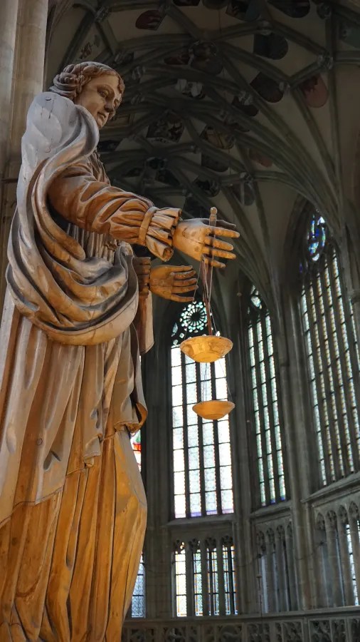 A wooden statue of Justice holding scales inside a gothic cathedral with tall windows
