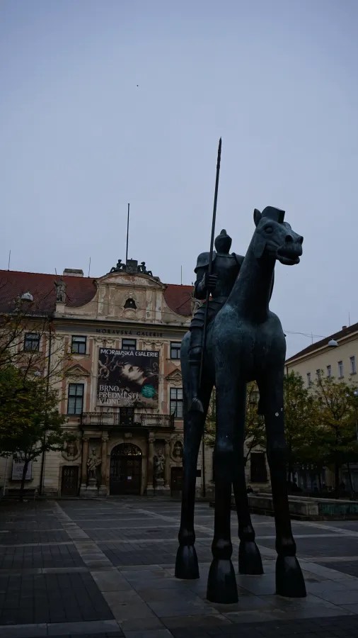 Tall equestrian statue of a knight with long legs in front of the Moravian Gallery building.