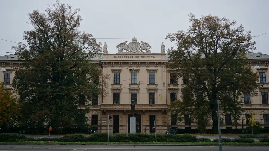 The beige facade of Masarykova Univerzita with a central sculpture and large flanking trees.
