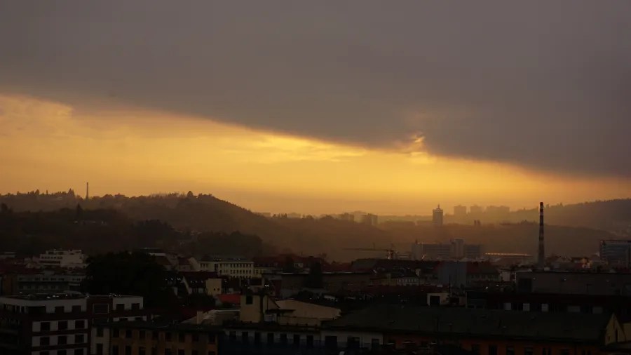 Golden sunset light breaking through dark clouds over a silhouetted cityscape and rolling hills
