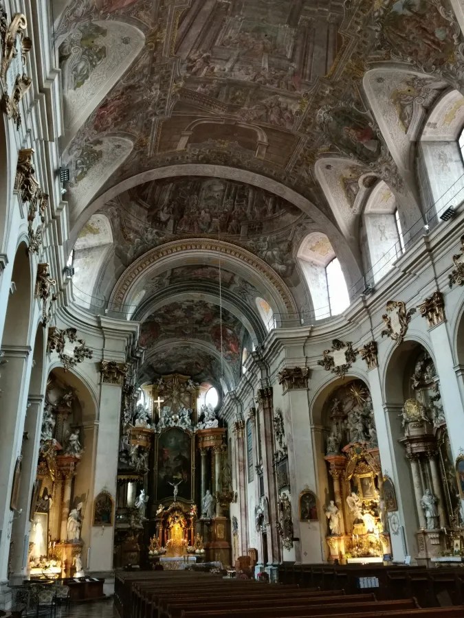 Ornate Baroque church interior with elaborate ceiling frescoes, gold-detailed altars, and rows of wooden pews.