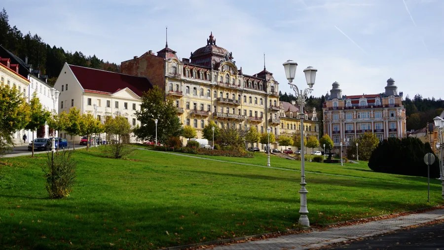 20221023_1_Tschechien_Marienbad (35) Ornate historic buildings and street lamps bordering a sloping green lawn in a town.