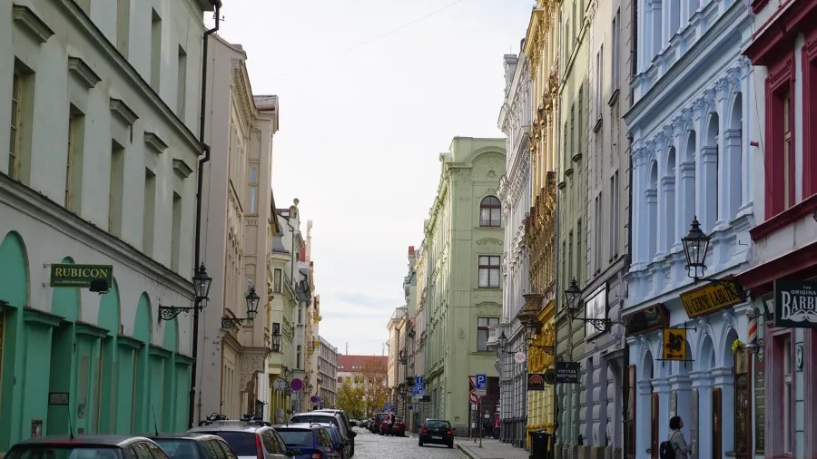 20221023_2_Tschechien_Pilsen (34) Narrow European street with colorful buildings, parked cars, and signs for Rubicon and U Černé Labutě.