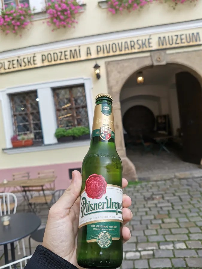 20221023_2_Tschechien_Pilsen (43) A hand holding a Pilsner Urquell beer bottle in front of the PLZEŇSKÉ PODZEMÍ A PIVOVARSKÉ MUZEUM.