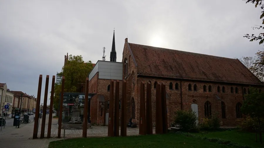 Historic brick building with a spire and modern weathered steel posts under an overcast sky.