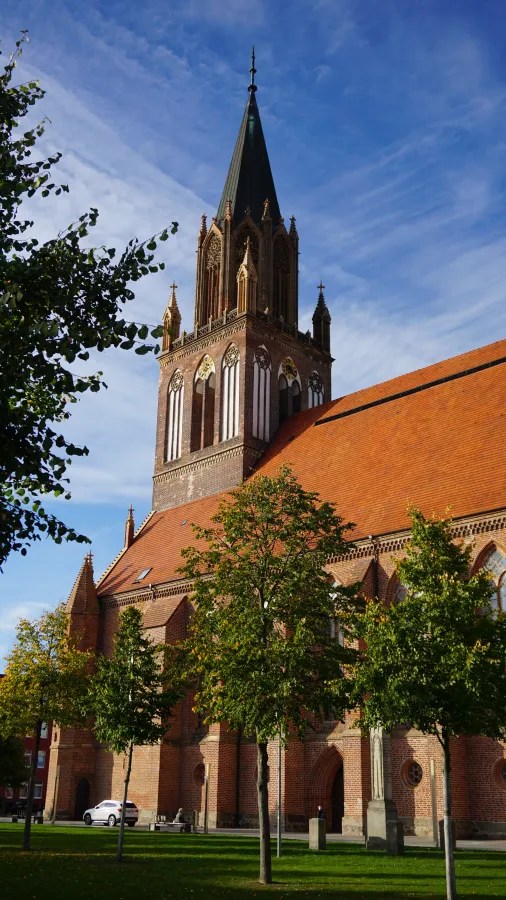 Red brick Gothic church with a tall spire and orange tiled roof behind green trees.