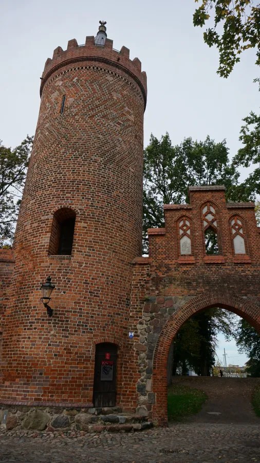 Medieval red brick tower and arched gatehouse surrounded by green trees under an overcast sky