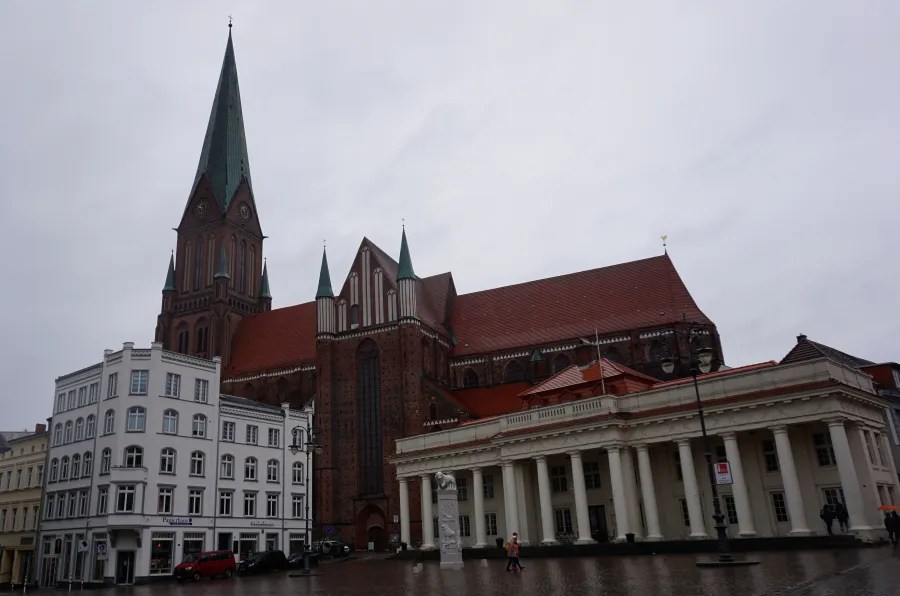 Large brick church with a tall green spire overlooking a square with classical buildings.